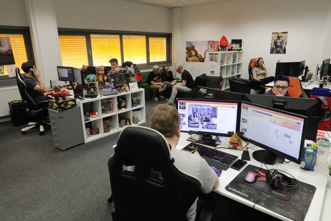 Freaks4UGaming office with people working at computers, decorated shelves with gaming items, and a small group collaborating in the back of the room.