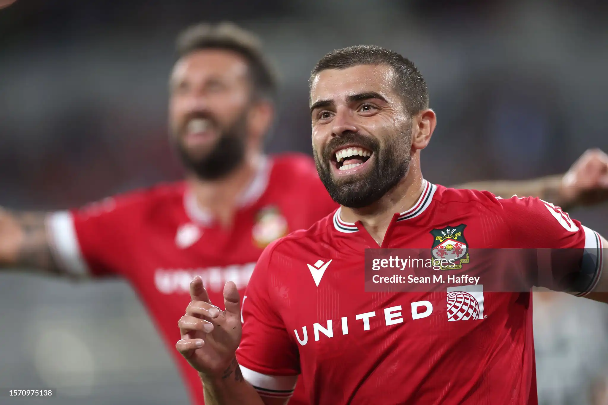 Wrexham AFC player celebrating a goal during a match, smiling while wearing the team’s red jersey with the club badge and sponsor logos visible.